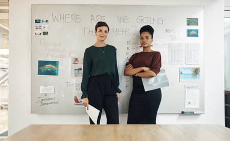 About to kill this presentation. Cropped portrait of two attractive young businesswomen standing in front of a whiteboard in their office boardroom.の写真素材