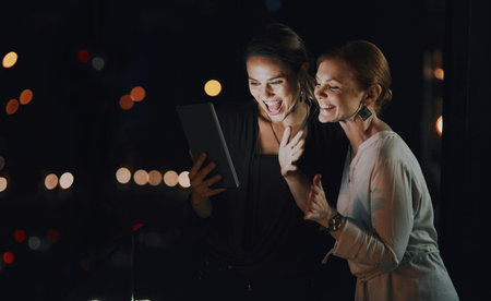 Connecting with global clients has never been easier. two businesswomen using a digital tablet to make a video call outside an office at night.の写真素材