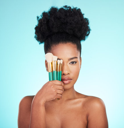Portrait, brushes and black woman with skincare, cosmetics and treatment against blue studio background. Face, African American female and lady with cosmetic tools, grooming and confidence with glowの写真素材