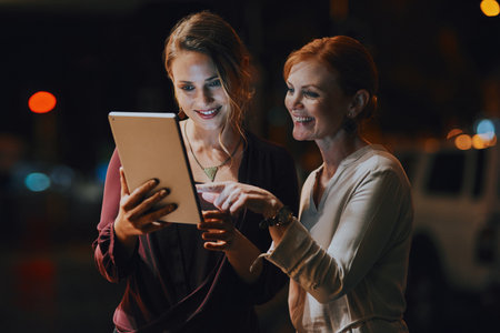 Devoted to getting the deadline done. two businesswomen using a digital tablet together outside an office at night.の写真素材