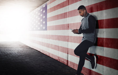Music, phone and black man runner relax against an American flag background during training run outdoors. Online, podcast and search by male athlete browse, chill and browse after workout in the USAの写真素材
