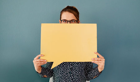 Heres my opinion on this...Studio portrait of an attractive young businesswoman covering her face with a speech bubble while sitting against a grey background.の写真素材