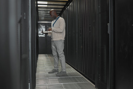 Server room, data center or IT black man with laptop for research, engineer working in dark server...