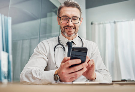 Healthcare, smile and doctor with smartphone at desk for wellness research, medical app and online consulting. Hospital, clinic and happy man with phone for internet, communication and telehealthの写真素材