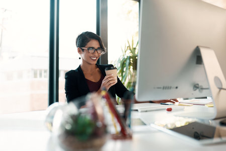 Coffee and work go hand in hand. an attractive young businesswoman sitting alone and holding a cup of coffee while using her computer.の写真素材