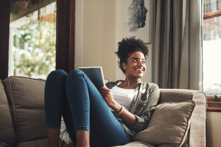 The weather is finally clearing up. a beautiful young woman using a digital tablet while relaxing on her sofa at home.の写真素材