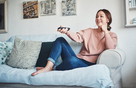 Looking for the perfect home movie. Full length shot of an attractive young woman sitting on her home sofa alone and using a television remote control.の写真素材