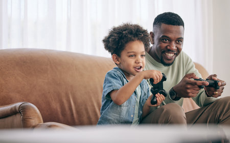 Black family, dad and child playing video games on living room sofa together with controllers at home. Happy African American father with son with smile enjoying bonding time on console entertainmentの写真素材