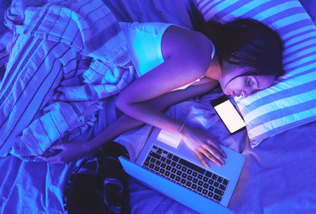 Woman, night and technology while sleeping in home bed with light from laptop and phone in bedroom. Tired student person with burnout asleep online for remote work or mockup social media chat on topの写真素材