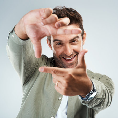 I can see this shot taking place. Portrait of a cheerful young man making frames with his hands to simulate the angle of a camera while standing against a grey background.の写真素材