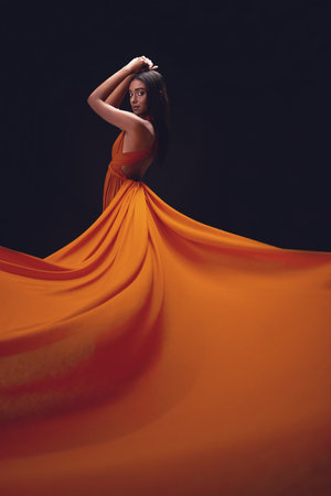 Fashion, woman and elegant with beauty on dark background, portrait and model in orange dress in studio. Indian female, glamour and stylish ballgown with sexy person, luxury style and designer wearの写真素材