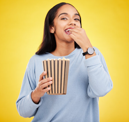 Happy woman is eating popcorn, watching tv and portrait, box with snack for movie on yellow studio background. Streaming service, film and food with corn treat, female with smile and cinemaの写真素材