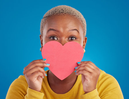 Paper, heart and shy with black woman in studio for love, date and kindness. Invitation, romance and feelings with female and shape isolated on blue background for emotion, support and affectionateの写真素材