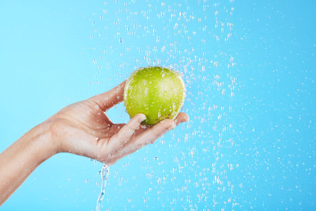 Water drops, apple cleaning and hands with fruit for wellness, healthcare and self care. Healthy food, green and natural skincare with a hand holding health snack for diet in blue background studioの写真素材