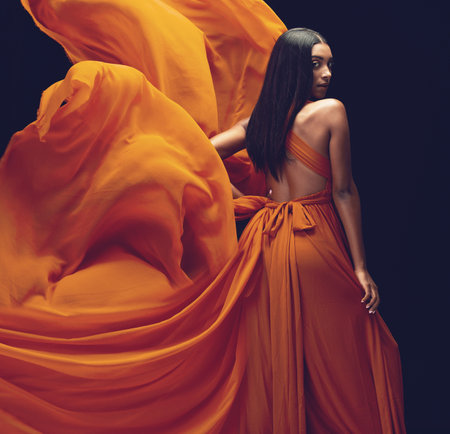 Fashion, portrait and woman in orange dress in studio isolated on a black background. Beauty, aesthetics and confident Indian female model with glamour, stylish and wind, trendy or designer ballgown.の写真素材
