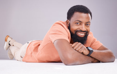 Portrait, happy and a black man lying on the floor isolated on a grey studio background. Smile, relax and a calm African guy relaxing, smiling and looking confident on the ground of a backdropの写真素材