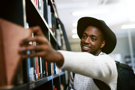 Fiction or non fiction, hes a fan of both. a happy young man removing a book from a shelf in a college library.の写真素材