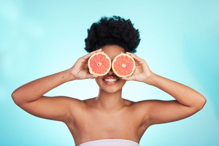 Black woman, face and smile with grapefruit for skincare nutrition, beauty or vitamin C against a blue studio background. Portrait of African female smiling with fruit for natural health and wellnessの写真素材