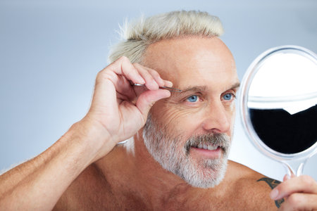 Senior man, tweezers and mirror check in a studio doing hair cleaning and grooming. Wellness, dermatology and beauty routine of a mature male model care with grey background and steel tools for faceの写真素材