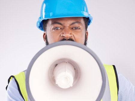 Engineering black man, megaphone and construction in studio portrait for angry shouting by white background. Engineer, architect or manager with loudspeaker in workplace with anger on frustrated faceの写真素材