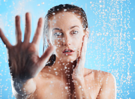 Shower portrait, water drops and woman hand doing skin cleaning, wellness and beauty routine in bathroom. Isolated, blue background and studio with young female doing hair care and dermatology facialの写真素材