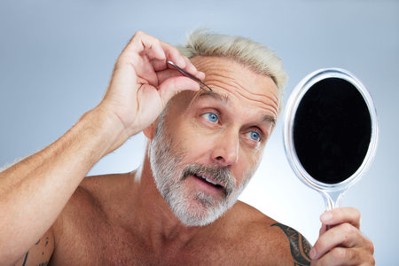 Senior man, tweezers and eyebrow in studio with mirror, grooming and self care for beauty by...