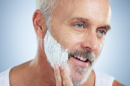 Mature man, face and shaving cream for grooming, skincare or hair removal against studio background. Closeup of senior male applying shave creme, cosmetics or product for haircare or facial treatmentの写真素材