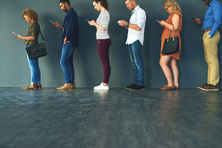 Getting busy with our phones. a group of people standing in a row behind each other while using their phones.の写真素材