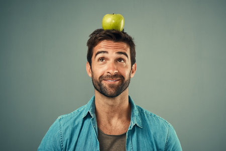 Your target should be a healthier lifestyle. Studio shot of a handsome young man posing with an apple on his head against a grey background.の写真素材