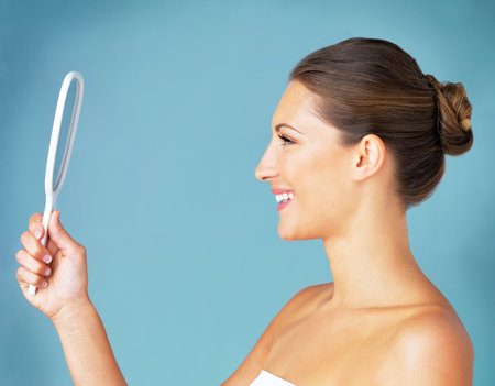 Its how you see yourself that counts. Studio shot of a beautiful young woman looking at herself in the mirror against a blue background.の写真素材