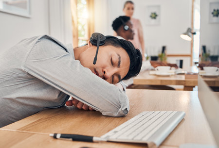 Tired call center worker sleeping at desk, burnout from working at telemarketing company and stress from consulting with people online on computer. Customer service employee with sleep problemの写真素材