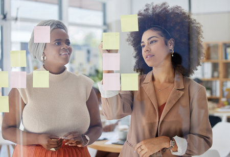 Team, brainstorming and business people with glass board, writing ideas on sticky note with collaboration and strategy. Planning, meeting and women working together, female employees and partnershipの写真素材