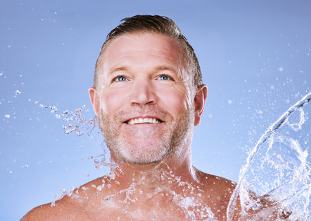 Wellness, water splash and senior man cleaning, morning routine and washing on blue studio background. Male, mature gentleman and daily hygiene with clear liquid for dermatology, skincare or groomingの写真素材