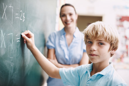 Extra lessons. A young boy doing mathematics on the blackboard as his teacher watches with a smile.の写真素材