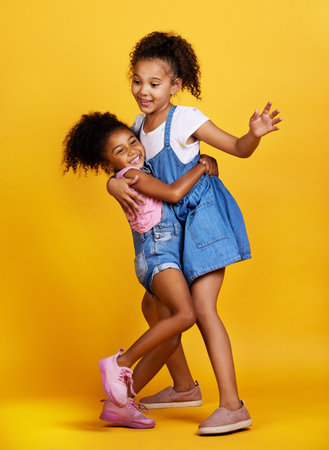 Children, hug and happy sisters in studio dancing with love, care and support of family on yellow background. Cute young girl kids play together for happiness, fun and positive attitude or fashionの写真素材