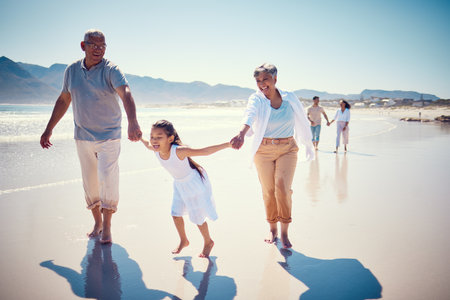 Beach, holding hands and playing, grandparents with girl, family walking on ocean sand together. Fun, vacation and happy senior man and woman with children bonding, quality time and summer in nature.の写真素材
