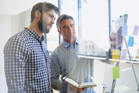 Their ideas are sure to please the client. two male coworkers arranging sticky notes on a glass wall...