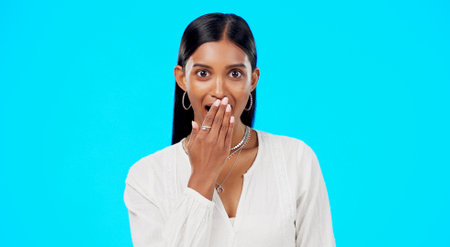 Woman, surprise and face with hand on mouth for comic story, secret or laugh for funny gossip by blue background. Young, gen z model and indian girl with laughing, amazed and wow for news in portraitの写真素材