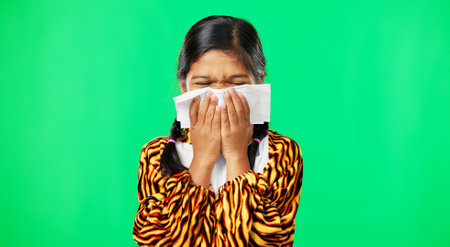Children, covid and blowing nose with a girl on a green screen background suffering from allergies. Portrait, kids and sick with a little female child sneezing from infection, pollen or hayfeverの写真素材