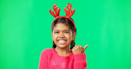 Christmas, green screen and a girl in a reindeer antlers headband pointing towards chromakey space. Portrait, kids and holidays with an adorable little female child on blank space feeling festiveの写真素材