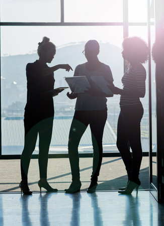 Late afternoon brainstorming session. Silhouette shot of female coworkers talking while standing in front of a window in an office.の写真素材