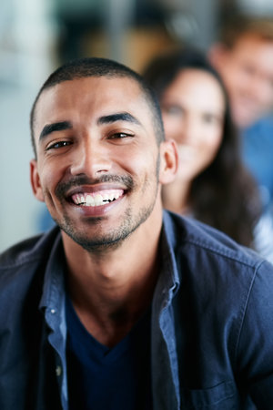 Doing what I love. Portrait of a smiling young professional sitting in an office with colleagues in the background.の写真素材