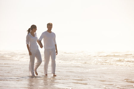 Another blissful day with someone special. Full length shot of an attractive young couple dressed in white walking along a beach.の写真素材