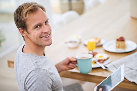 Carefree morning at home. Portrait of a handsome man using his tablet while having breakfast at home.の写真素材