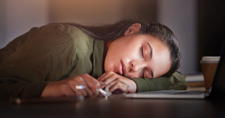 Tired, business woman and sleeping in office at night with burnout, stress problem and low energy in evening. Fatigue, lazy and overtime female employee nap at table, deadlines and rest of overworkedの写真素材