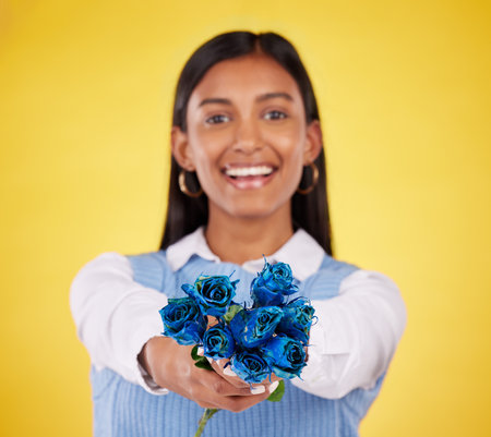 Portrait, love and roses with a woman on a yellow background in studio for valentines day. Face, blue flowers or smile with a happy young female holding a plant for romance or anniversary celebrationの写真素材