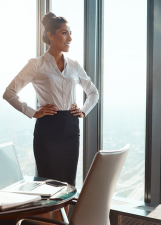 Enjoying the view from the top. an attractive young businesswoman standing with her hands on her hips in the office.の写真素材