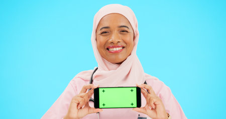 Phone, green screen and woman doctor isolated on blue background healthcare, tracking marker and mockup space. Face of nurse or muslim person with medical mobile app for advertising video in studioの写真素材