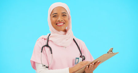 Woman, doctor and clipboard portrait in studio for information or medical records. Islamic or muslim female and professional nurse in hijab with smile and document for healthcare on blue backgroundの写真素材