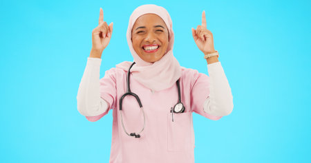 Muslim nurse, happy woman and pointing finger portrait with space for healthcare, religion or happiness. Islamic female medical worker with hand, smile and hijab for announcement on blue backgroundの写真素材
